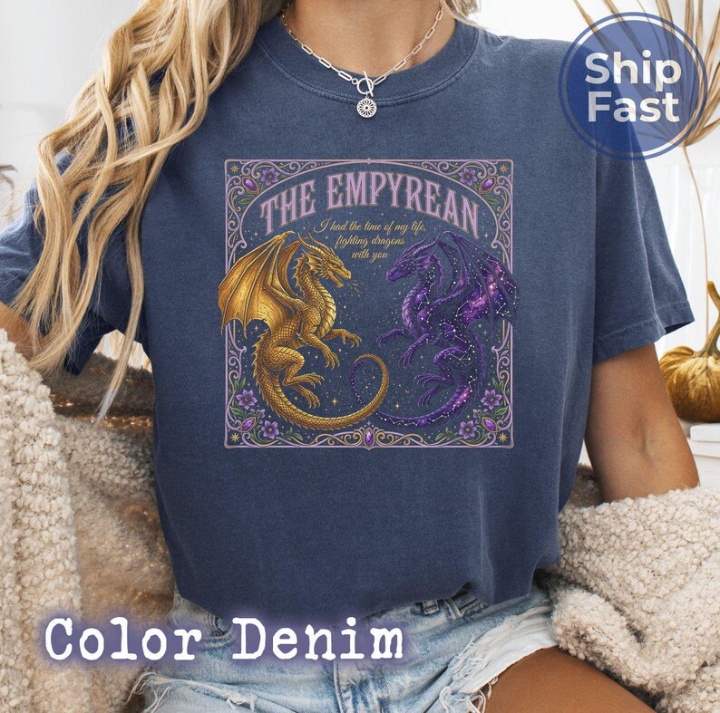 May include: A denim blue t-shirt featuring a detailed graphic of two dragons, one gold and one purple, facing each other. The text "THE EMPYREAN" and a quote are above the dragons. The shirt is a relaxed fit.