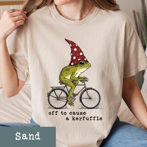 May include: Sand-colored t-shirt featuring a whimsical illustration of a green frog wearing a red and white polka-dotted hat, riding a bicycle. The text "off to cause a kerfuffle" is printed below the image. The shirt is made of soft material.