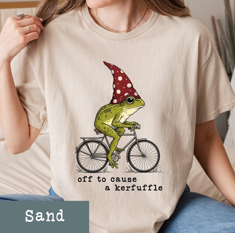 May include: Sand-colored t-shirt featuring a whimsical illustration of a green frog wearing a red and white polka-dotted hat, riding a bicycle. The text "off to cause a kerfuffle" is printed below the image. The shirt is made of soft material.