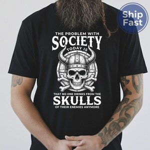 Puede incluir: Camiseta negra con un dise&ntilde;o gr&aacute;fico que presenta una calavera con un casco con cuernos, huesos cruzados y el texto "THE PROBLEM WITH SOCIETY TODAY IS THAT NO ONE DRINKS FROM THE SKULLS OF THEIR ENEMIES ANYMORE."