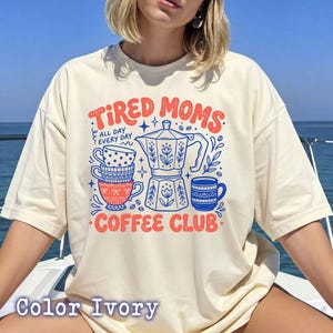 Könnte beinhalten: Elfenbeinfarbenes T-Shirt mit dem Text "TIRED MOMS COFFEE CLUB" in Rot und Blau. Das Design umfasst eine Kaffeekanne, gestapelte Tassen und dekorative Elemente. Das Shirt hat eine lockere Passform.