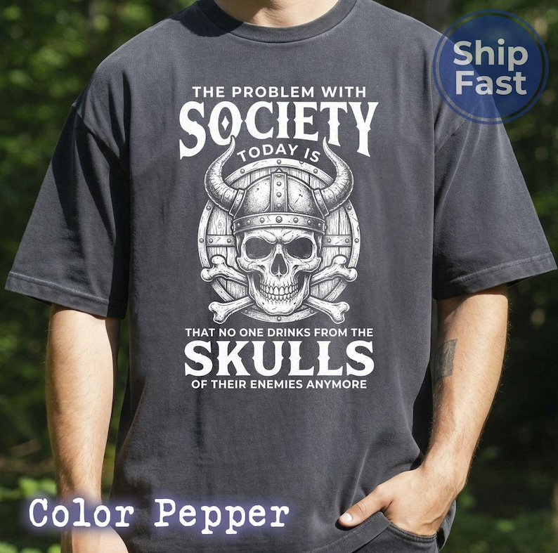Puede incluir: Camiseta color pimienta con un gr&aacute;fico que presenta una calavera con un casco con cuernos, huesos cruzados y el texto "THE PROBLEM WITH SOCIETY TODAY IS THAT NO ONE DRINKS FROM THE SKULLS OF THEIR ENEMIES ANYMORE."