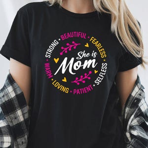 Może przedstawiać: Czarna koszulka z okrągłym wzorem z napisem "She is Mom" w białym skrypcie. Projekt zawiera słowa "Strong, Beautiful, Fearless, Selfless, Patient, Loving, Warm" w różowo-żółtej kolorystyce.