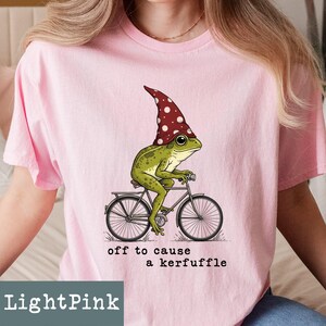 May include: Light pink t-shirt featuring a whimsical illustration of a green frog wearing a red and white polka-dot hat, riding a bicycle. The text "off to cause a kerfuffle" is printed below the graphic. The shirt is a light pink color.
