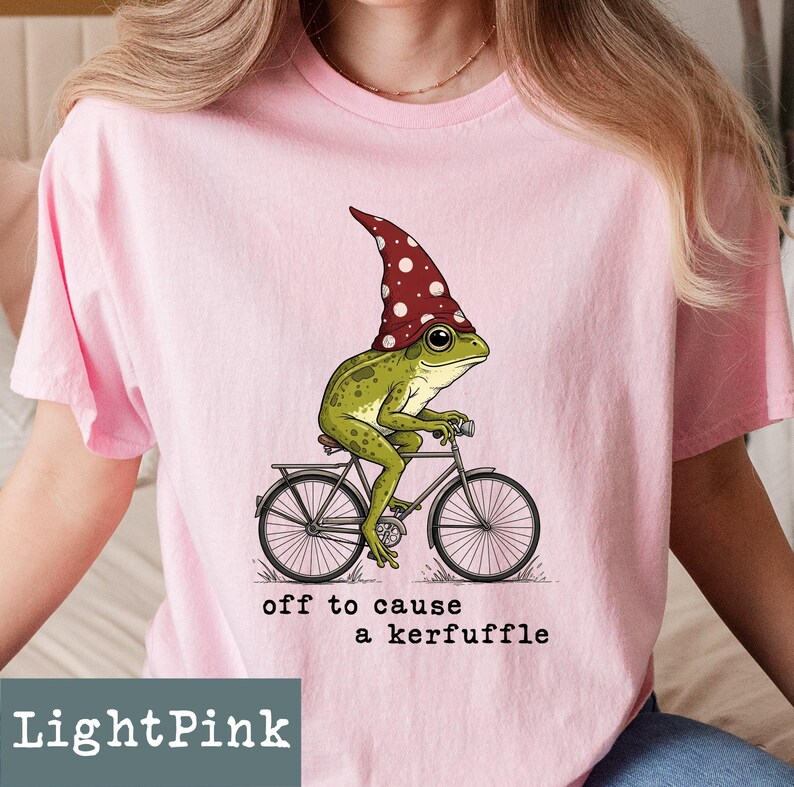 May include: Light pink t-shirt featuring a whimsical illustration of a green frog wearing a red and white polka-dot hat, riding a bicycle. The text "off to cause a kerfuffle" is printed below the graphic. The shirt is a light pink color.