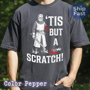 May include: Dark gray t-shirt with a graphic of a knight holding a sword, with the text "'TIS BUT A SCRATCH!". The knight has a bloody arm and a severed arm on the ground. The shirt is a relaxed fit.
