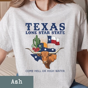 May include: Ash-colored t-shirt featuring a graphic of the Texas state flag, a longhorn, and a cactus. The text reads "TEXAS LONE STAR STATE" and "COME HELL OR HIGH WATER".