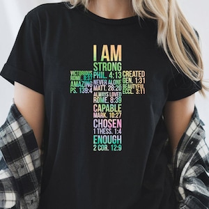 May include: Black t-shirt featuring a cross-shaped design with the words "I AM STRONG" and various inspirational phrases and Bible verses. The shirt has a crew neck and short sleeves.