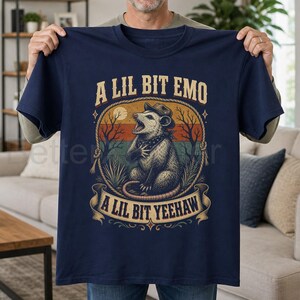 May include: A navy blue short-sleeve t-shirt featuring a graphic of an opossum wearing a cowboy hat and bandana. The design includes a sunset background, a rope border, and the text A LIL BIT EMO and A LIL BIT YEEHAW in a western font.