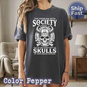 Puede incluir: Camiseta gris oscuro con un gr&aacute;fico blanco de una calavera con un casco con cuernos, con el texto "THE PROBLEM WITH SOCIETY TODAY IS THAT NO ONE DRINKS FROM THE SKULLS OF THEIR ENEMIES ANYMORE."