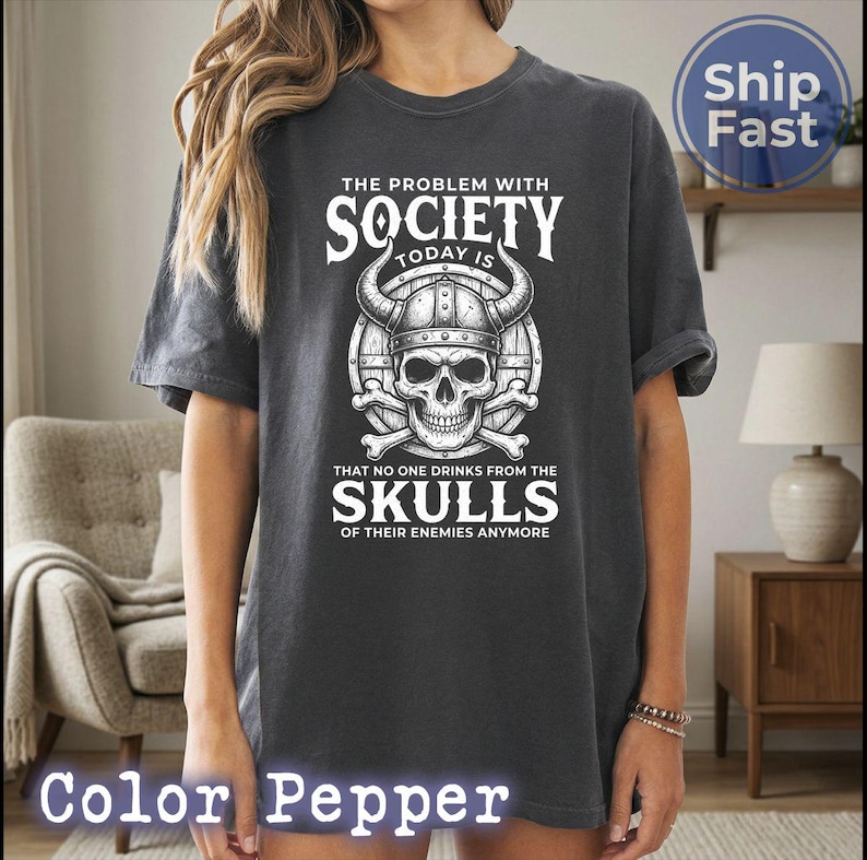 Puede incluir: Camiseta gris oscuro con un gr&aacute;fico blanco de una calavera con un casco con cuernos, con el texto "THE PROBLEM WITH SOCIETY TODAY IS THAT NO ONE DRINKS FROM THE SKULLS OF THEIR ENEMIES ANYMORE."