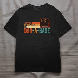 May include: Black t-shirt featuring the text "I keep all my dad jokes in a Dad-a-base" in orange, yellow, and teal. A retro floppy disk graphic is to the right of the text. The shirt has short sleeves and a crew neck.