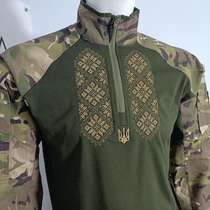 May include: A green and camouflage long-sleeved shirt with a half-zip. It has gold embroidered floral patterns on the chest and a gold trident emblem. The shoulders and collar are patterned with camouflage.