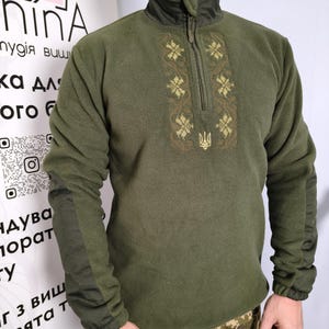 Op de afbeelding: Olijfgroene fleece trui met een hoge kraag en een kwartrits. De voorkant heeft gouden geborduurde bloemen en een Oekraïense drietand. De mouwen hebben donkerdere groene panelen. Camouflage broek.