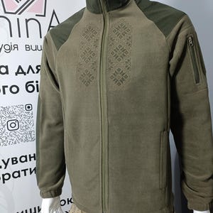 Puede incluir: Chaqueta de forro polar verde oliva con cuello alto y cremallera completa. La chaqueta presenta parches oscuros en los hombros y un patrón bordado decorativo en la parte delantera. Un pequeño bolsillo con cremallera es visible en la manga izquierda.