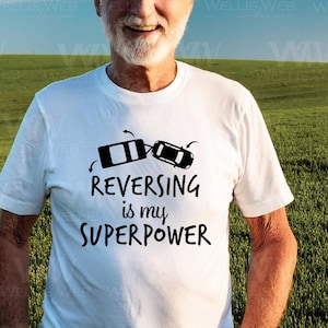 May include: White t-shirt with the text "REVERSING is my SUPERPOWER" in black. The graphic includes two black car illustrations with arrows. The shirt is a casual wear item.