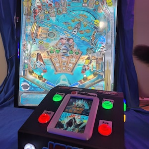 May include: A Jaws-themed pinball machine with a black cabinet and a playfield featuring shark and ocean graphics. The machine has a digital display and buttons labeled "Launch", "Fire", and "Quit". The Jaws logo is prominently displayed.