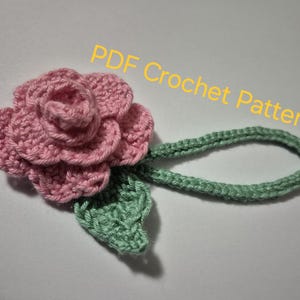 Puede incluir: Una rosa de crochet con hojas verdes y un tallo. La rosa es rosa y las hojas son de color verde claro. El tallo es de color verde claro y está tejido en punto de crochet simple. El texto "PDF Crochet Pattern" está escrito en letras amarillas sobre un fondo blanco.