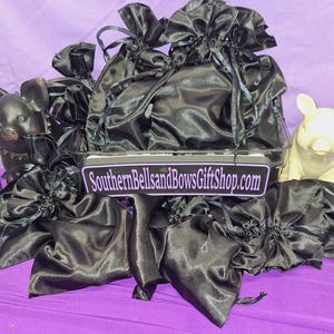 May include: A collection of black satin drawstring bags, some tied with ribbons, arranged in a clear container. The bags are on a purple surface, with a sign that reads "SouthernBellsandBowsGiftShop.com". Decorative pig figurines are on either side.