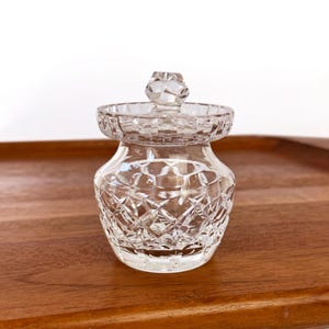 May include: A clear, cut-glass jar with a lid. The jar has a diamond pattern and a small, decorative knob on top. The jar is sitting on a wooden tray.