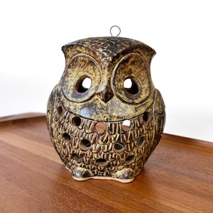 May include: A ceramic owl-shaped candle holder in shades of brown and tan. The owl has large eye holes and a series of smaller holes around the body. A metal loop is attached to the top for hanging. The candle holder is sitting on a wooden tray.