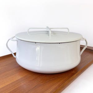 May include: A light gray enamelware pot with a matching lid. The pot has two side handles and a lid handle. The pot is sitting on a wooden tray. The pot is a light gray color with a black trim.