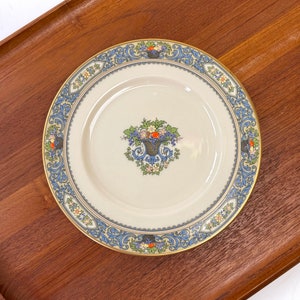 May include: A white porcelain dinner plate with a gold rim and a blue and gold floral design. The center of the plate features a basket of fruit with green leaves and red berries.