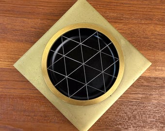 May include: A square gold-colored base supports a circular black tray featuring a white geometric grid pattern of intersecting lines. The object is composed of a flat, diamond-oriented gold plate with a centered, raised circular black insert.