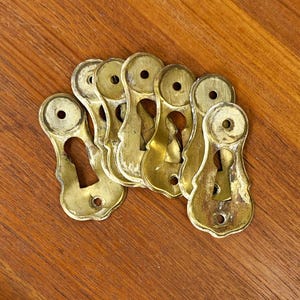 May include: A collection of six brass keyhole escutcheon plates, each featuring a vertical teardrop-shaped opening for a key and two small circular holes at the top and bottom for mounting screws, displaying a metallic gold finish with patina.