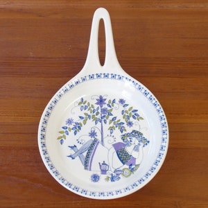 May include: A white ceramic serving dish with a blue and green floral design. The dish features a woman in a purple dress sitting under a tree with a teapot and flowers.