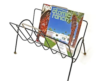 May include: Black metal magazine rack with a curved design. The rack is holding two magazines, one with the title "atomic ranch mid century marvels" and the other with the title "food & cook".