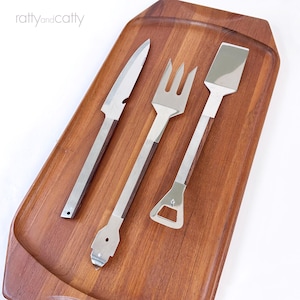May include: A set of stainless steel grilling tools with wooden handles, including a knife, fork, and spatula, arranged on a rectangular wooden serving tray. The tray has a recessed area. The text "ratty and catty" is in the upper left corner.