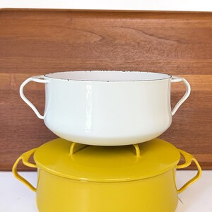 May include: A white enamel pot with two side handles sits atop a mustard yellow enamel pot with a matching lid and two side handles. The white pot features a dark rim, and both vessels have a smooth, rounded shape with curved metal handles.