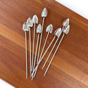 May include: Set of ten silver cocktail picks with heart-shaped ends. The picks are arranged on a wooden surface.