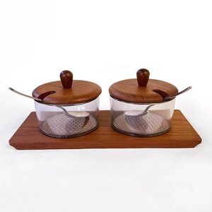 May include: A set of two clear glass spice jars with wooden lids and small spoons, set on a rectangular wooden tray. The lids have a dark brown knob on top. The spoons are silver. The tray and lids are a similar shade of brown.
