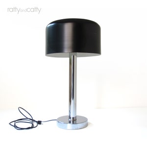 May include: A black and chrome table lamp with a cylindrical base and a round, black lampshade. The lamp has a cord and plug.
