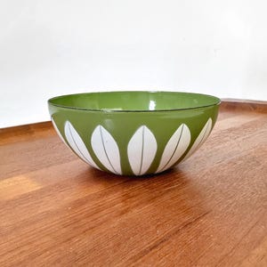May include: A round bowl with a smooth olive green exterior featuring a repeating pattern of white, petal-shaped motifs around the base. The interior of the bowl is a lighter shade of green, and the rim is finished with a thin, dark green line.