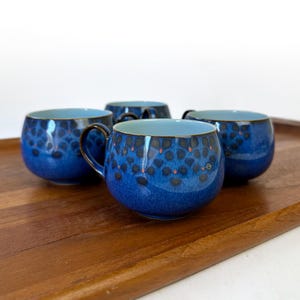 May include: Four blue ceramic mugs with handles, arranged on a wooden tray. The mugs have a speckled pattern in black and orange, with a lighter blue interior. The mugs are round and have a glossy finish.