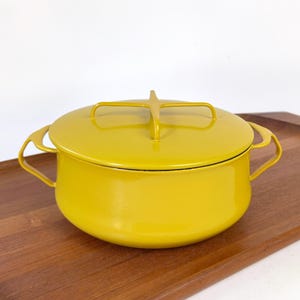 May include: A mustard yellow enameled metal casserole dish with a matching lid. The lid features a distinctive cross-shaped handle, and the pot has two curved side handles. The design is smooth and rounded, showcasing a vibrant solid color finish.