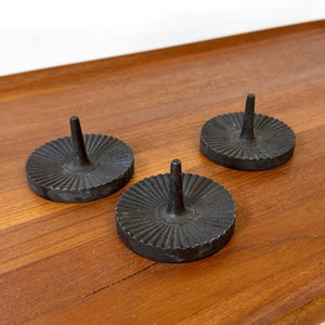 May include: Three black metal candle holders with a circular base and a central spike. The bases have a ridged, sunburst design. They are displayed on a wooden surface.