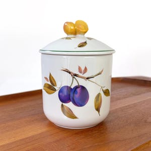 May include: White ceramic jar with a lid, featuring a plum branch design. The jar has a green rim and a lid with a yellow plum-shaped handle. The plums are purple, and the leaves are brown and yellow.