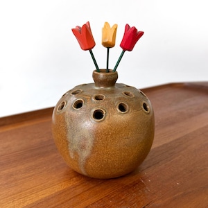 May include: A brown ceramic vase with multiple holes, holding three wooden tulip flowers. The tulips are painted red, yellow, and red, with green stems. The vase sits on a wooden surface.