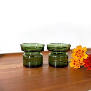 May include: Two green glass candle holders with a wide, round top and a narrow base. The candle holders are sitting on a wooden surface.