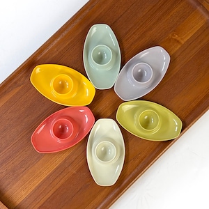 May include: Six colorful egg cups in various pastel shades, including yellow, green, red, and white, arranged in a flower shape on a wooden surface.
