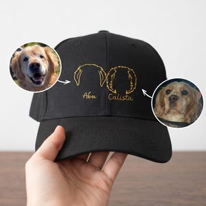 May include: A black baseball cap featuring two gold-colored line art dog silhouettes. Below the silhouettes, the names Abu and Calista are embroidered in gold. Two circular photo inserts show the dogs corresponding to the line art designs.