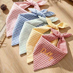 May include: A set of five triangular pet bandanas featuring a gingham check pattern in pink, blue, light green, yellow, and red. The red bandana is embroidered with the name Dolly in gold thread. Each bandana has tied ends for securing.