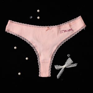 May include: Pink thong underwear with a ribbed texture, delicate lace trim, and a small bow. The word "Stella" is embroidered in a cursive font. The background is black with scattered pearls and a white lace bow.