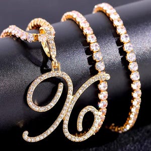 Peut inclure: Ensemble collier et bracelet de couleur or. Le collier pr&eacute;sente un pendentif en forme de lettre "X" en cursive incrust&eacute; de cristaux transparents. Le bracelet et la cha&icirc;ne du collier sont &eacute;galement orn&eacute;s de cristaux transparents, cr&eacute;ant un effet scintillant. Les bijoux sont pos&eacute;s sur un fond sombre.