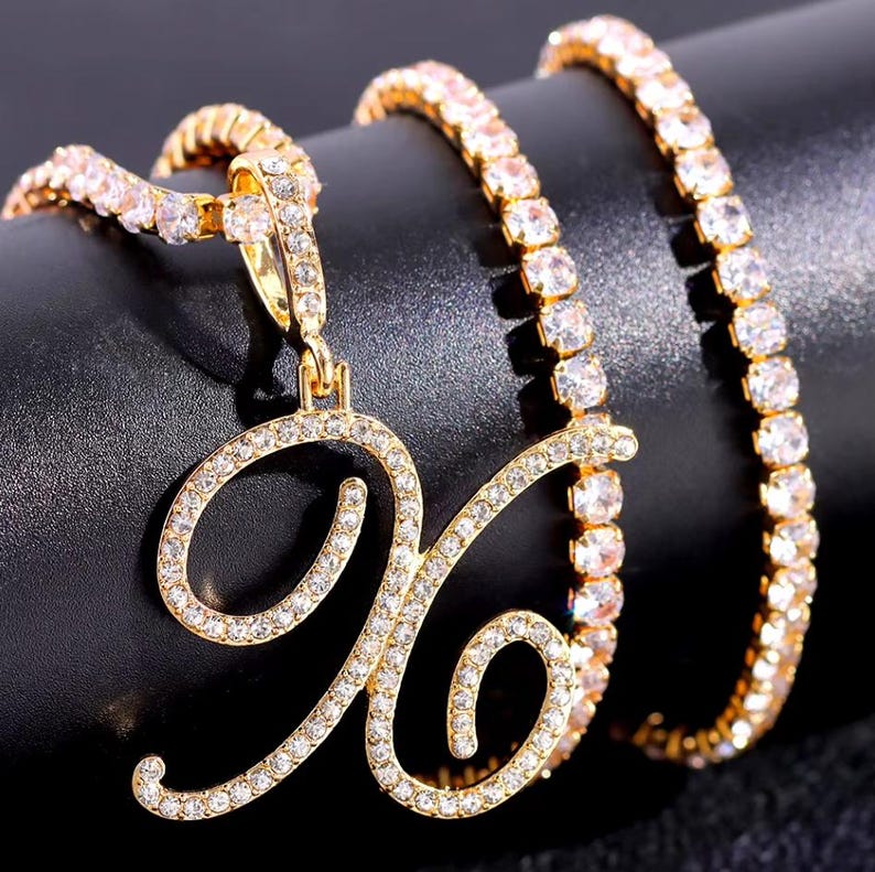 Peut inclure: Ensemble collier et bracelet de couleur or. Le collier pr&eacute;sente un pendentif en forme de lettre "X" en cursive incrust&eacute; de cristaux transparents. Le bracelet et la cha&icirc;ne du collier sont &eacute;galement orn&eacute;s de cristaux transparents, cr&eacute;ant un effet scintillant. Les bijoux sont pos&eacute;s sur un fond sombre.