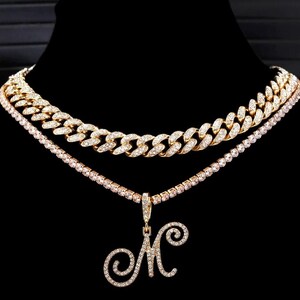 Peut inclure: Ensemble de colliers dor&eacute;s &agrave; trois niveaux. La couche sup&eacute;rieure est une cha&icirc;ne &eacute;paisse orn&eacute;e de diamants. La couche interm&eacute;diaire est un d&eacute;licat collier de tennis. La couche inf&eacute;rieure pr&eacute;sente un pendentif avec la lettre "M" incrust&eacute;e de diamants. Le fond est noir.